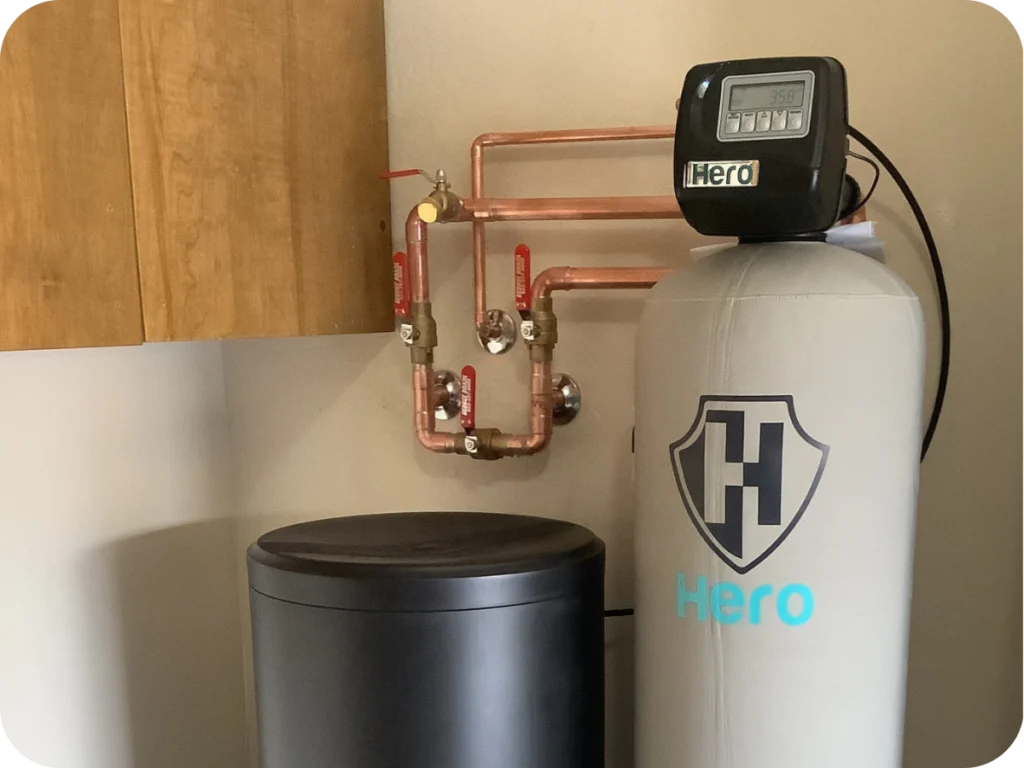 A water softener system labeled "Hero" is installed next to a black container and copper pipes, mounted on a beige wall near a wooden cabinet.
