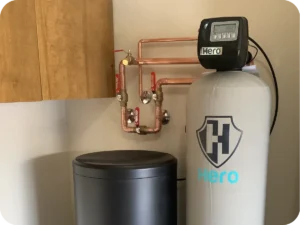 A Water Softener System Labeled &Quot;Hero&Quot; Is Installed Next To A Black Container And Copper Pipes, Mounted On A Beige Wall Near A Wooden Cabinet.
